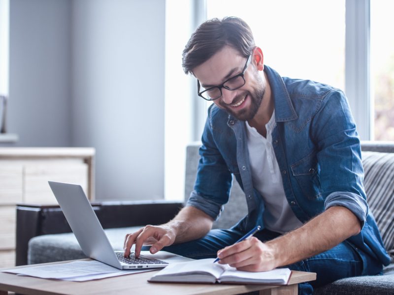 Handsome businessman in eyeglasses is making notes and smiling while working with a laptop at home